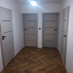 J.FELIŃSKI OKNA DRZWI ROLETY USŁUGI REMONTOWE JANUSZ FELIŃSKI - Korytarz po remoncie z trzema szarymi drzwiami z czarnymi klamkami i parkietem ułożonym w jodełkę. Centralnie widoczny włącznik światła.