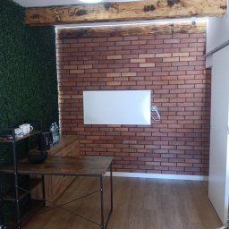 J.FELIŃSKI OKNA DRZWI ROLETY USŁUGI REMONTOWE JANUSZ FELIŃSKI - Pokój z ceglaną ścianą, drewnianymi belkami i zieloną ścianą z roślin. Widoczny stół z metalową konstrukcją i drewnianym blatem. Nowoczesny design z elementami rustykalnymi.