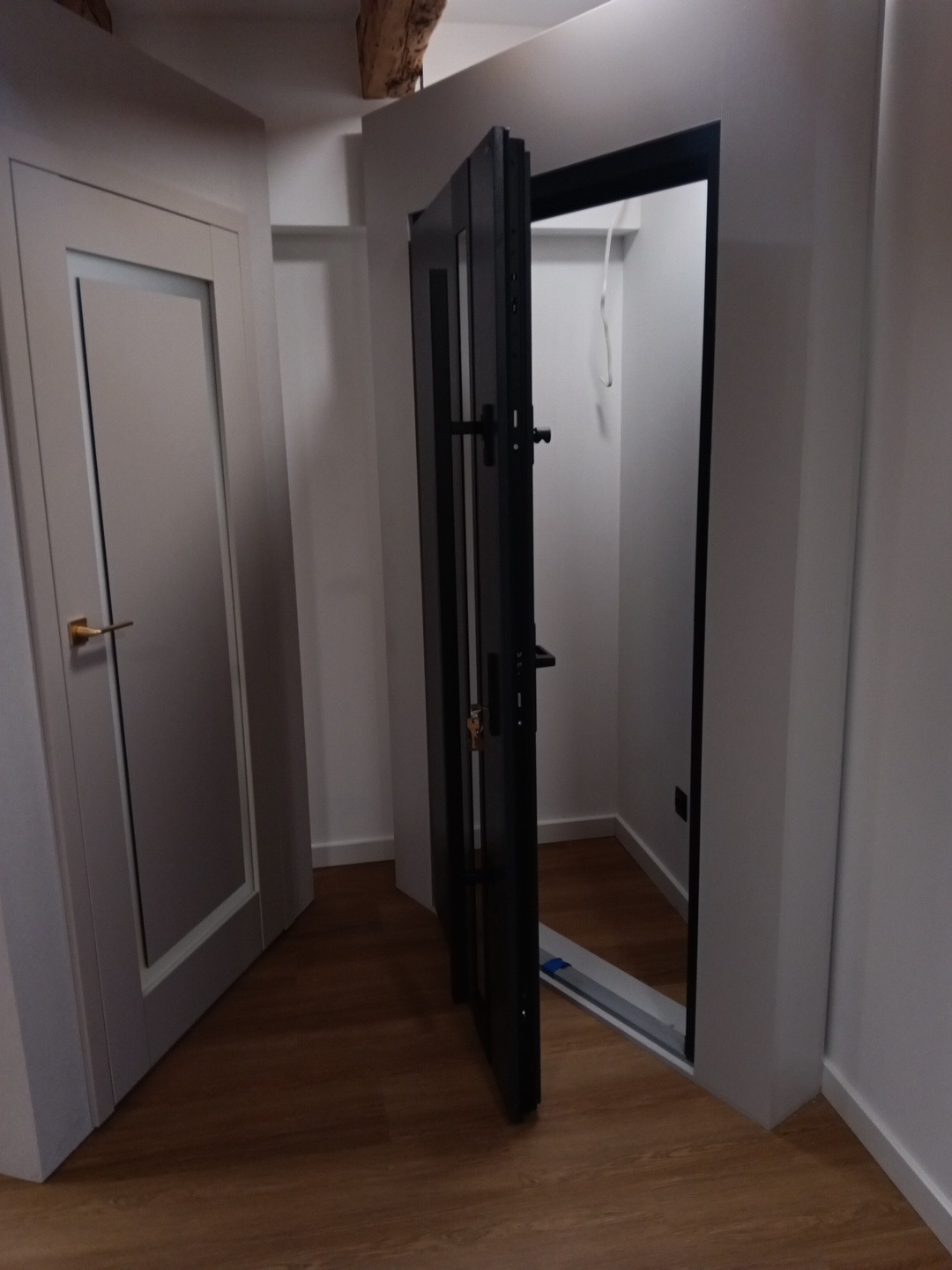 Ciemne, nowoczesne drzwi otwarte do garderoby, widoczne na tle drewnianej podłogi i białych ścian. Obok drzwi z przeszkleniem i złotą klamką. Widoczny fragment drewnianej belki stropowej.