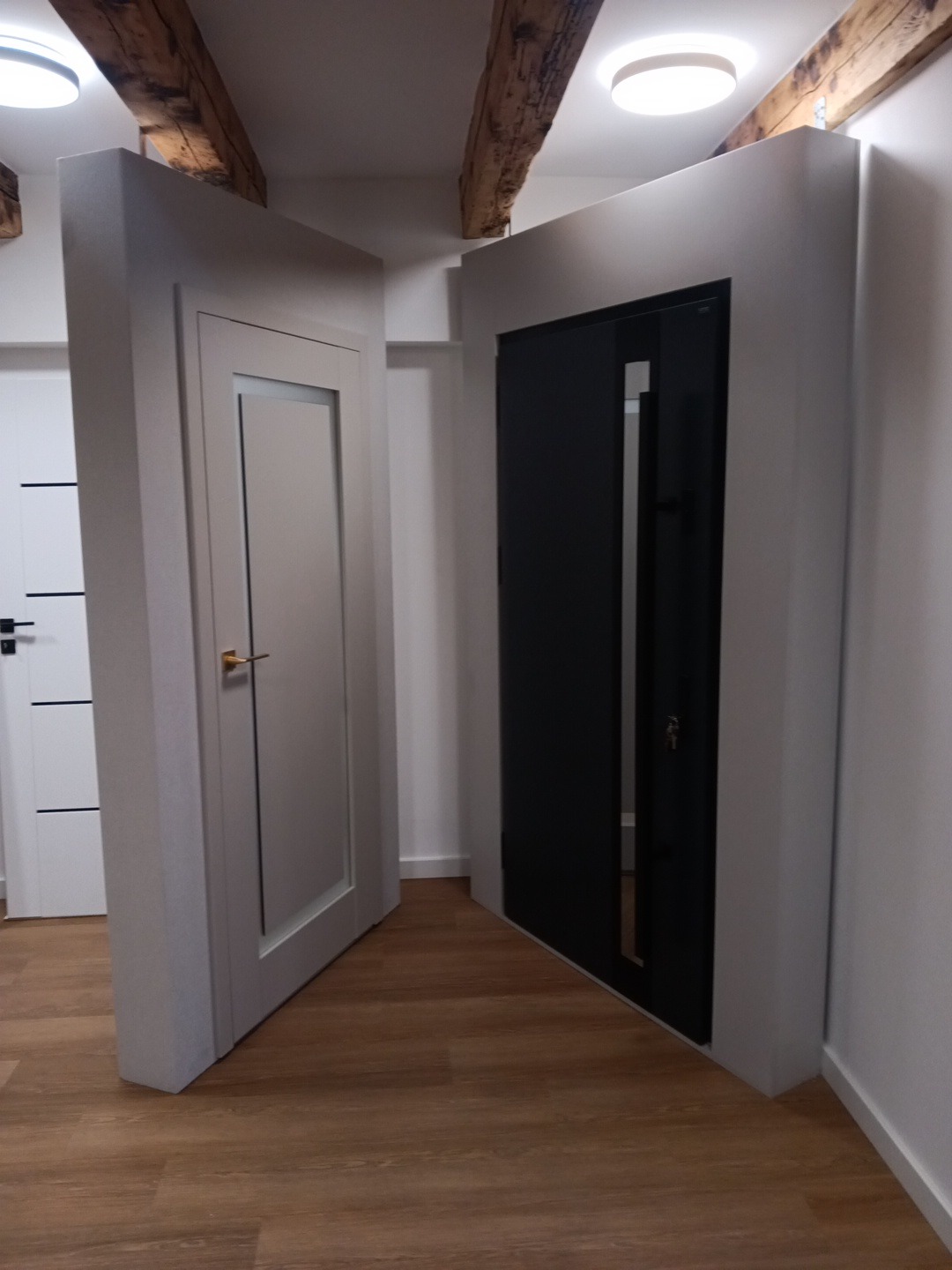 Dwa rodzaje drzwi w korytarzu: białe drzwi wewnętrzne z klamką i czarne drzwi wejściowe z zamkiem, drewniana podłoga, sufit z drewnianymi belkami i oświetleniem.