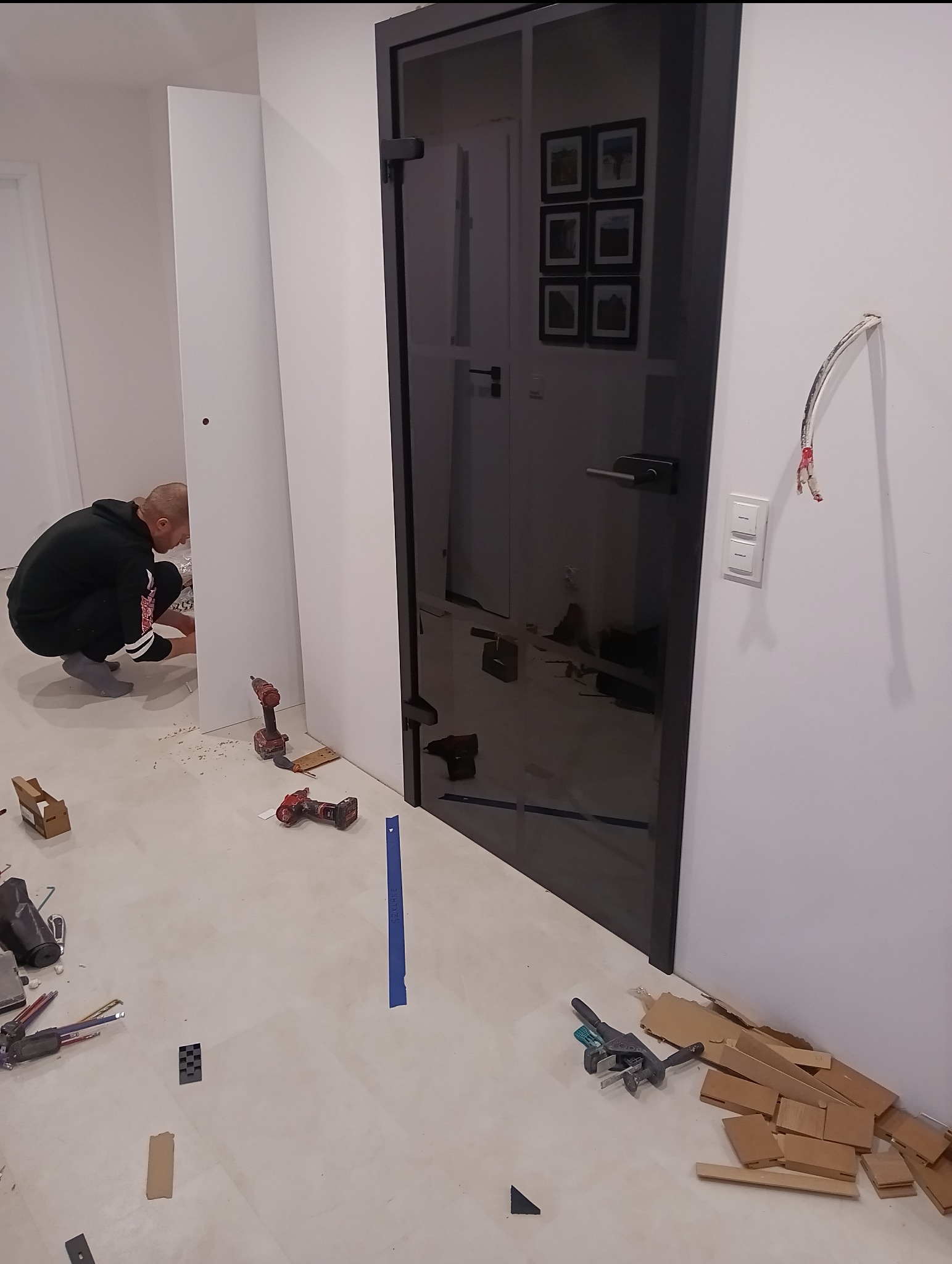 Montaż czarnych, szklanych drzwi wewnętrznych. Widoczne narzędzia, w tle osoba przy białej ścianie. Na podłodze resztki materiałów i akcesoriów montażowych.