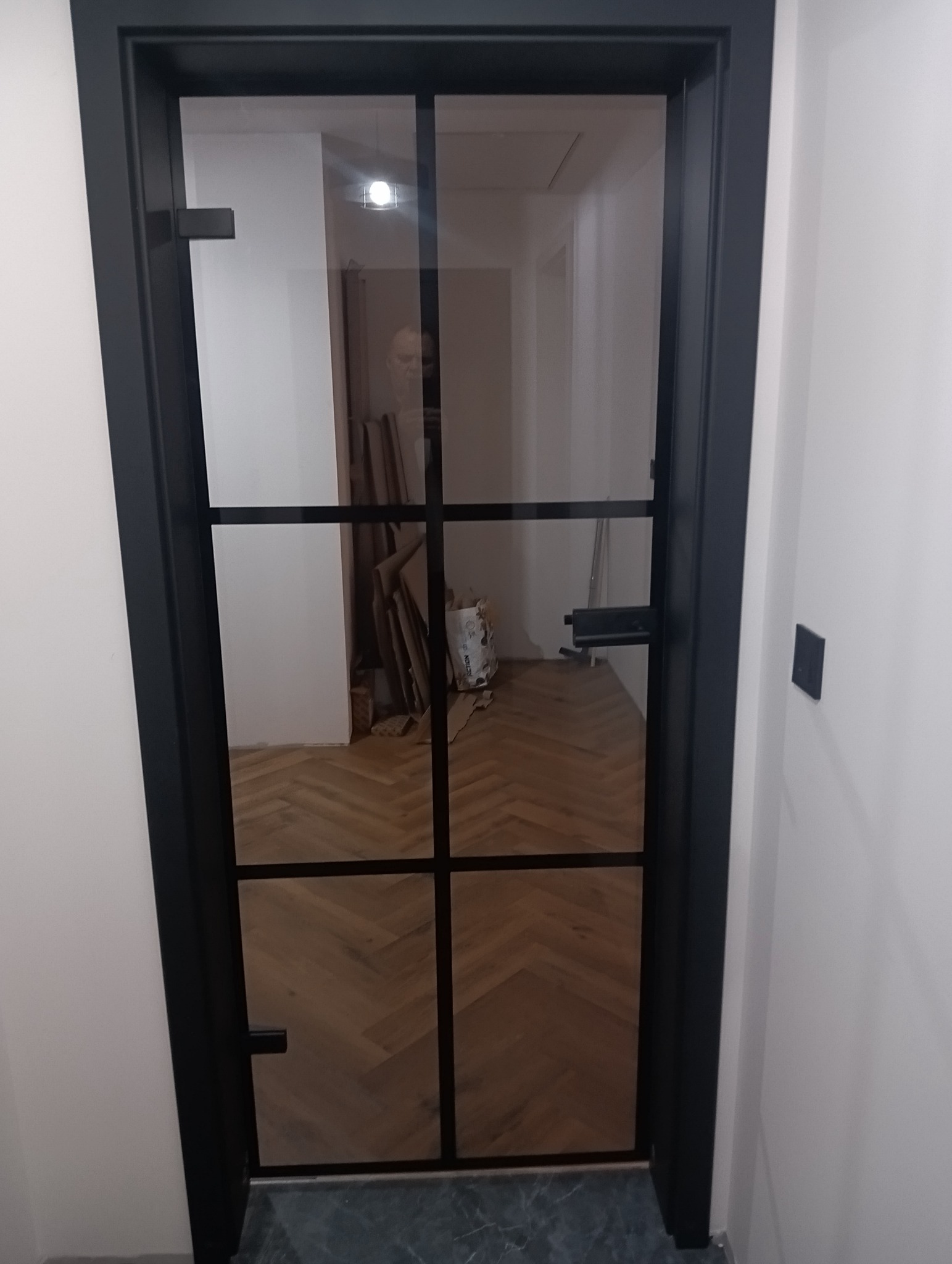 Nowoczesne drzwi wewnętrzne z czarną ramą i szklanym wypełnieniem, widok na parkiet ułożony w jodełkę. Minimalistyczny design, jasne ściany.