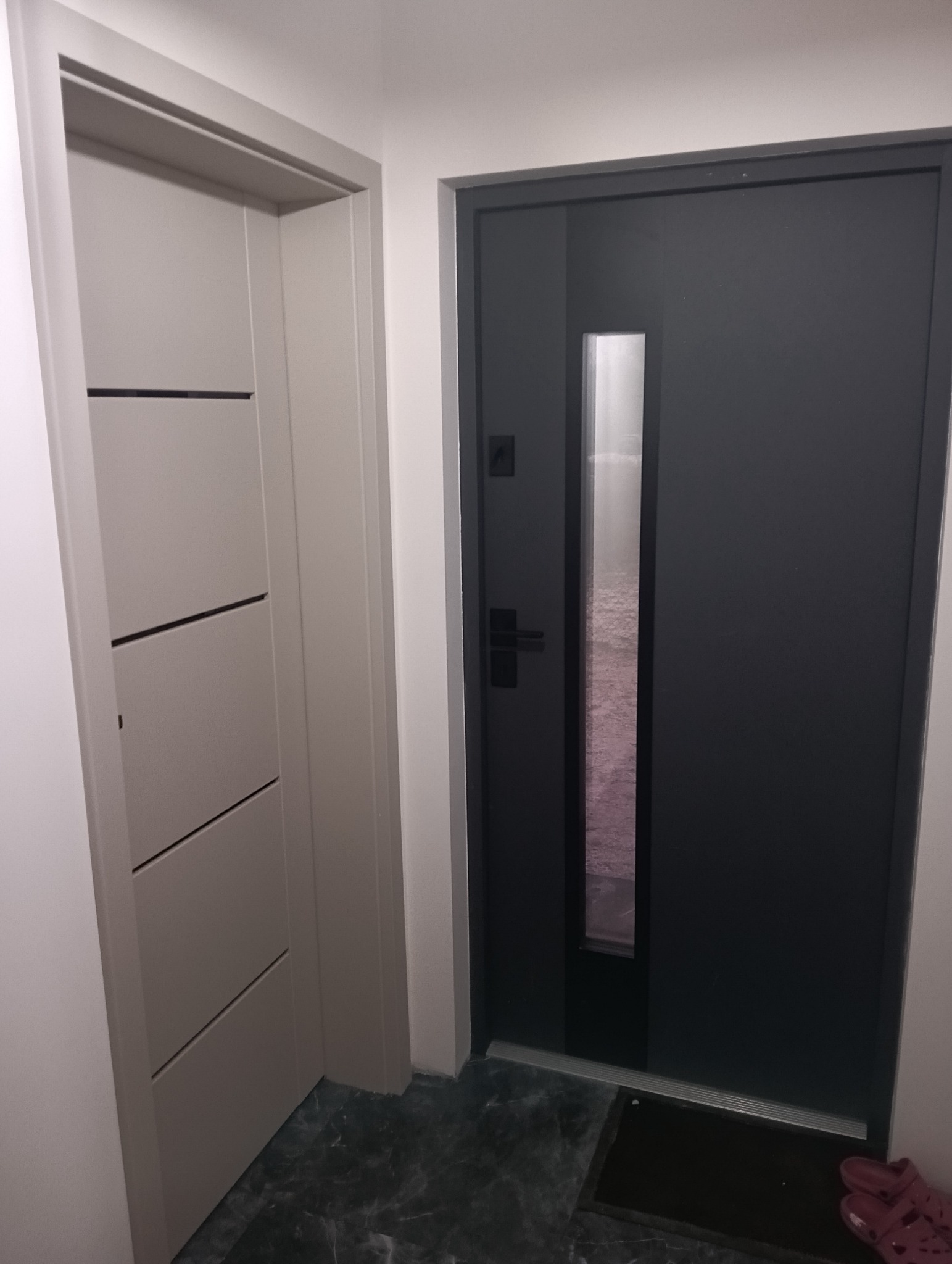 Nowoczesne, grafitowe drzwi antywłamaniowe z szybą, widoczne obok drzwi wewnętrznych z poziomymi frezami. Na podłodze ciemny marmur.