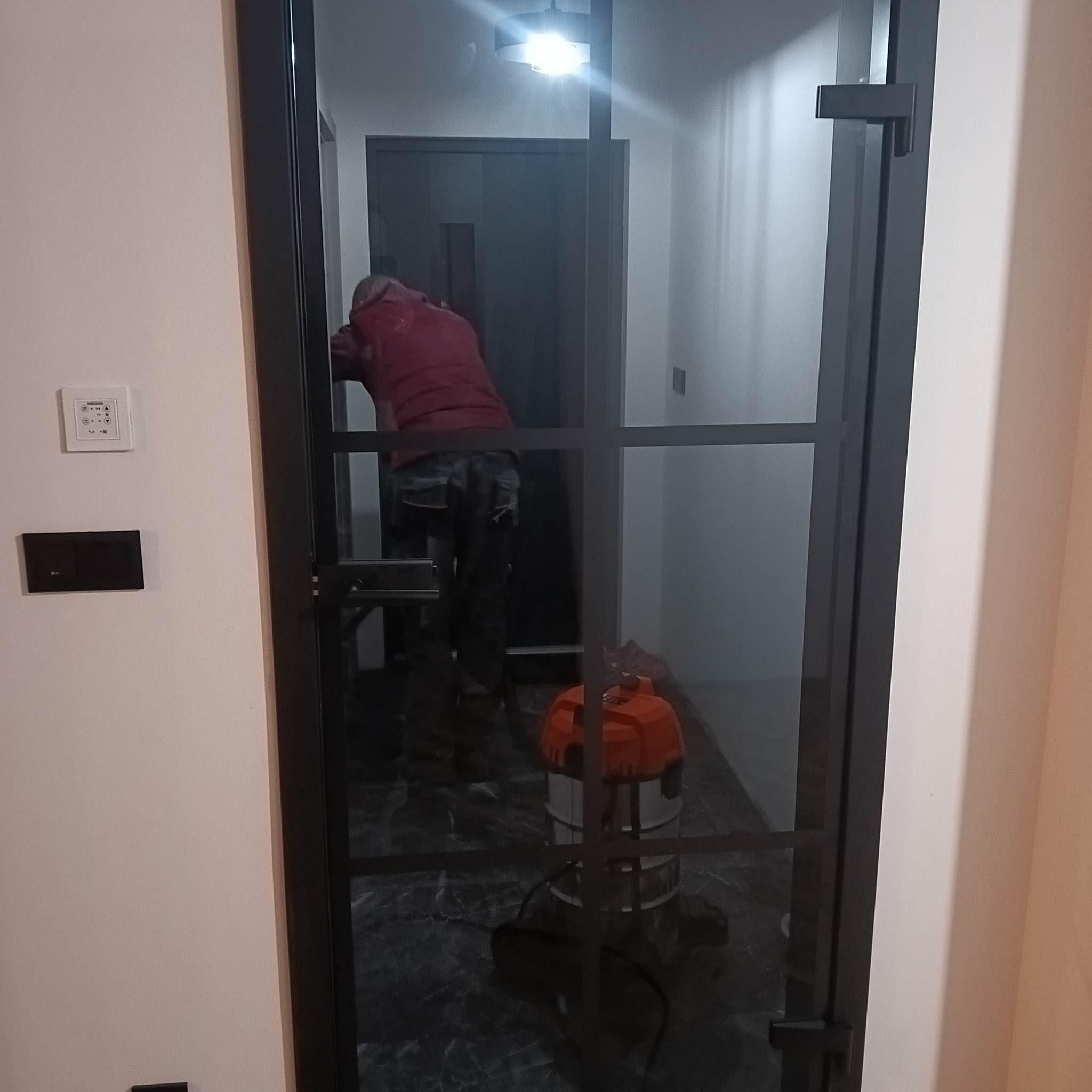Nowoczesne, czarne drzwi z szybami w metalowej ramie, widoczne w korytarzu. W tle osoba i odkurzacz. Marmurowa podłoga odbija światło.