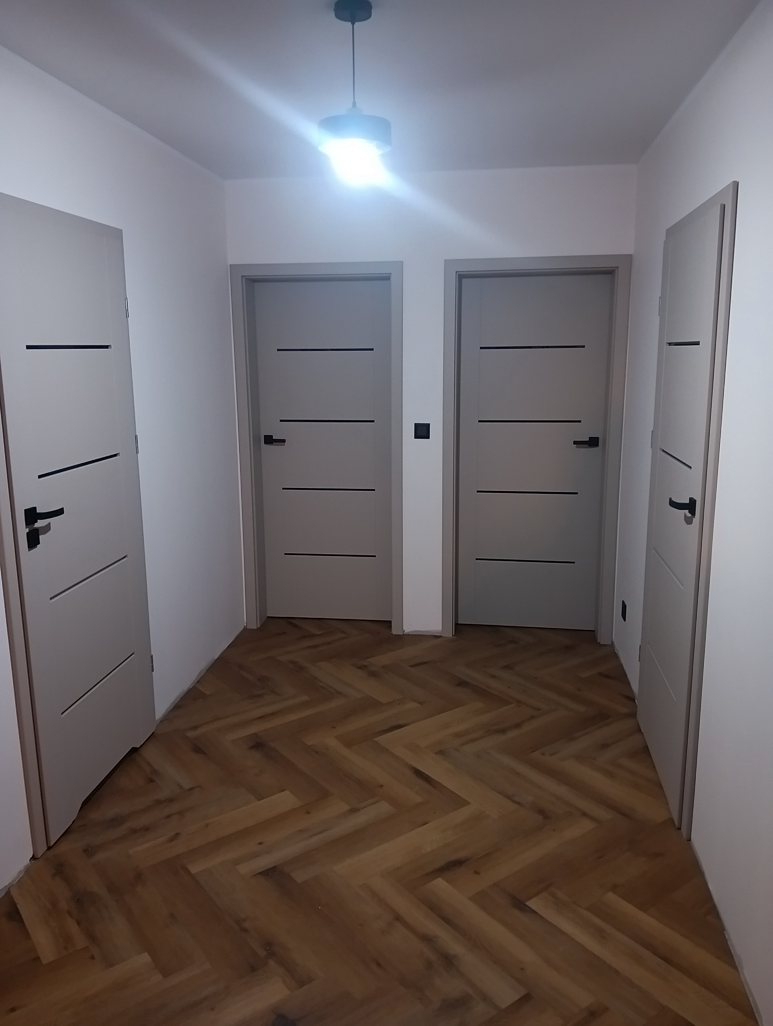Korytarz po remoncie z trzema szarymi drzwiami z czarnymi klamkami i parkietem ułożonym w jodełkę. Centralnie widoczny włącznik światła.