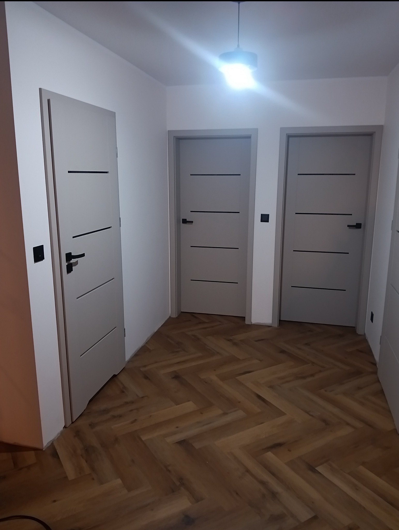 Jasne, nowoczesne drzwi wewnętrzne z czarnymi akcentami w korytarzu z parkietem w jodełkę. Widok na trzy skrzydła drzwiowe i oświetlenie sufitowe.