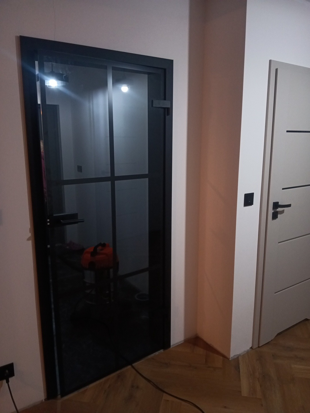 Ciemna, szklana, wewnętrzna rama drzwiowa z widocznym narzędziem w tle, zamontowana w jasnym pomieszczeniu obok białych drzwi z poziomymi frezami.