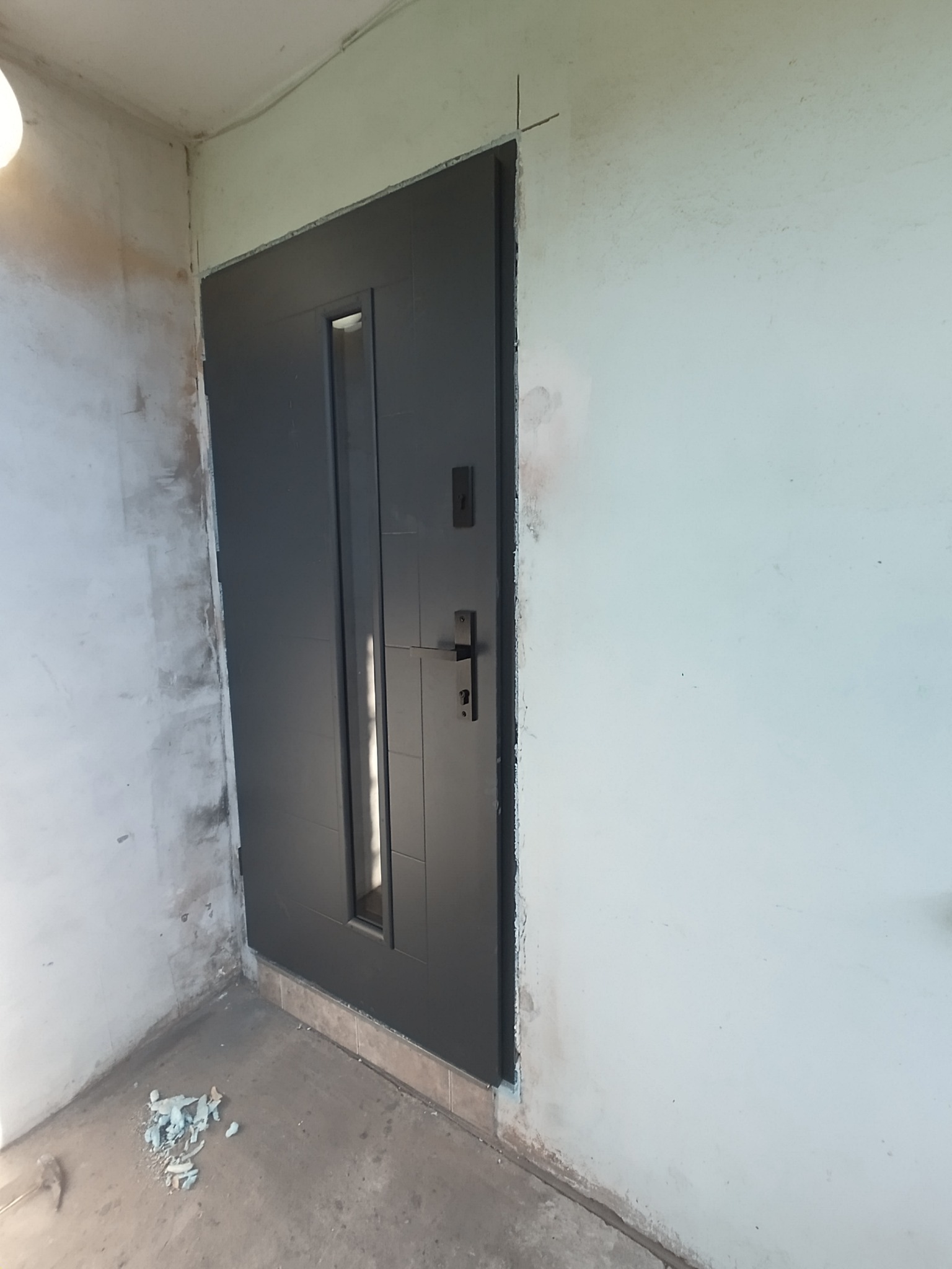 Zamontowane, grafitowe drzwi wejściowe z szybą, widoczne w surowym otworze. Ściany w trakcie wykończenia, widoczne ślady po pracach budowlanych.