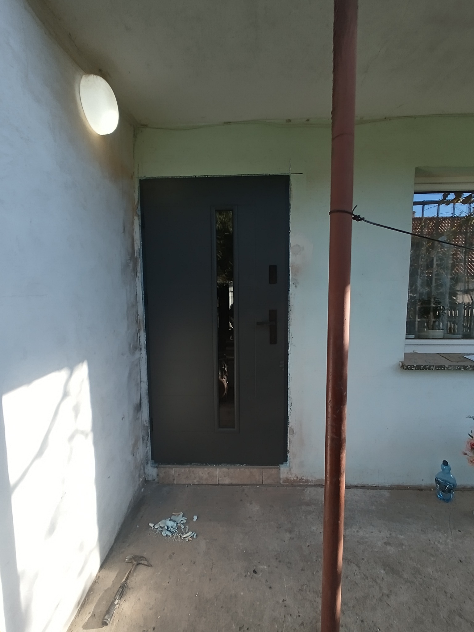 Zamontowane, grafitowe drzwi wejściowe z szybą, widoczne od zewnątrz. Na podłodze leżą resztki gruzu i narzędzie. Ściany wymagają wykończenia.