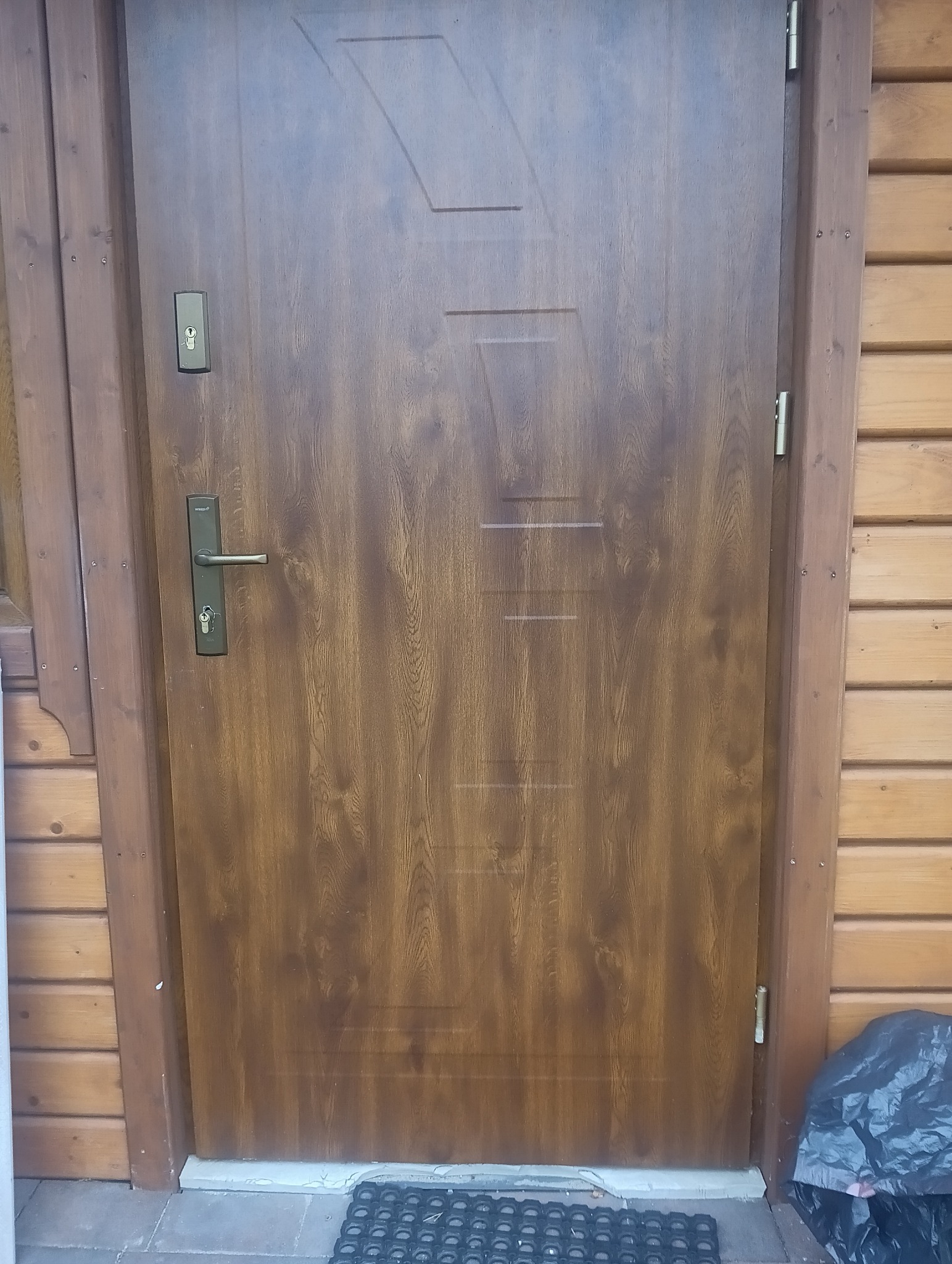 Brązowe drzwi antywłamaniowe z drewnianą ościeżnicą, widoczne od frontu, z zamontowanym zamkiem i klamką. Obok ściana z drewnianych paneli.