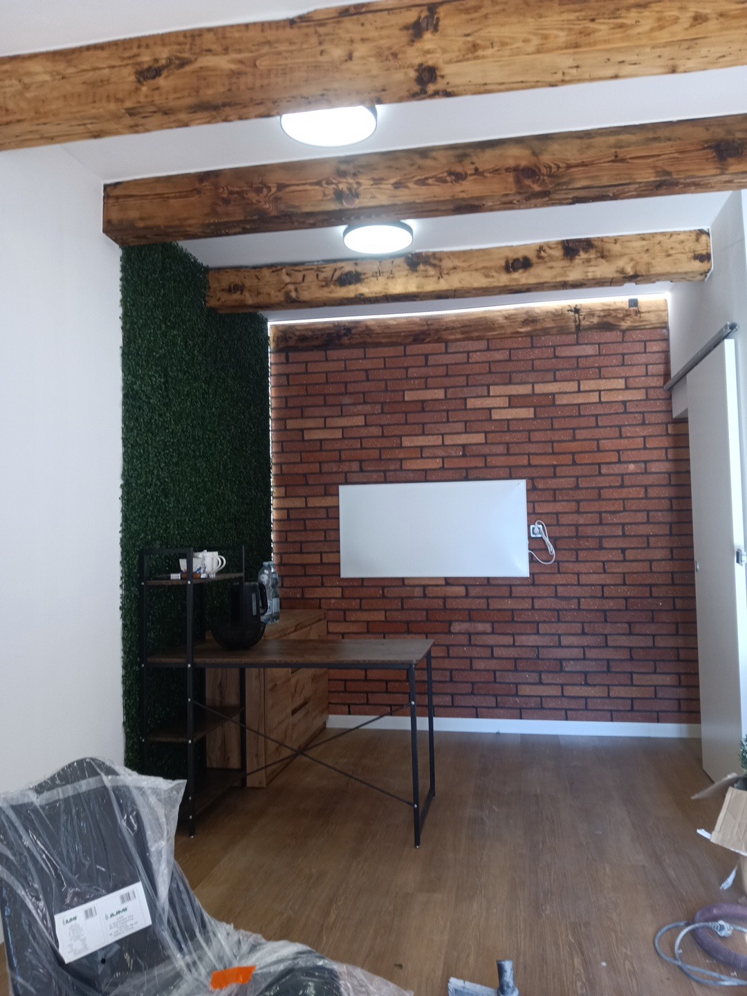Nowoczesne wnętrze z ceglaną ścianą, drewnianymi belkami stropowymi i zieloną ścianą z roślin. Widoczny fragment biurka i regału. Podłoga drewniana.