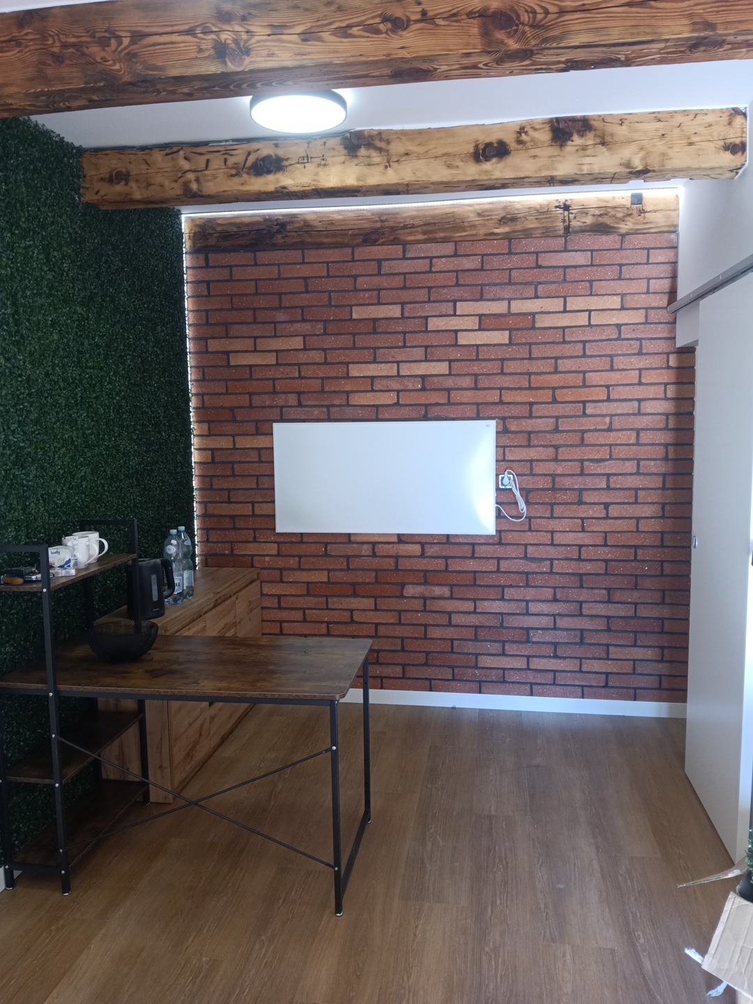 Pokój z ceglaną ścianą, drewnianymi belkami i zieloną ścianą z roślin. Widoczny stół z metalową konstrukcją i drewnianym blatem. Nowoczesny design z elementami rustykalnymi.