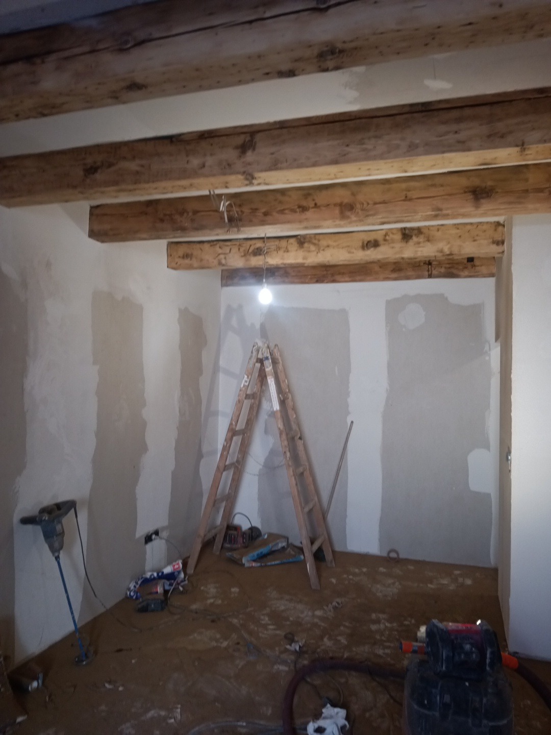 Wnętrze pokoju w trakcie prac remontowych: drewniane belki stropowe, ściany częściowo pokryte gładzią, drabina, narzędzia malarskie i sprzęt budowlany na podłodze.