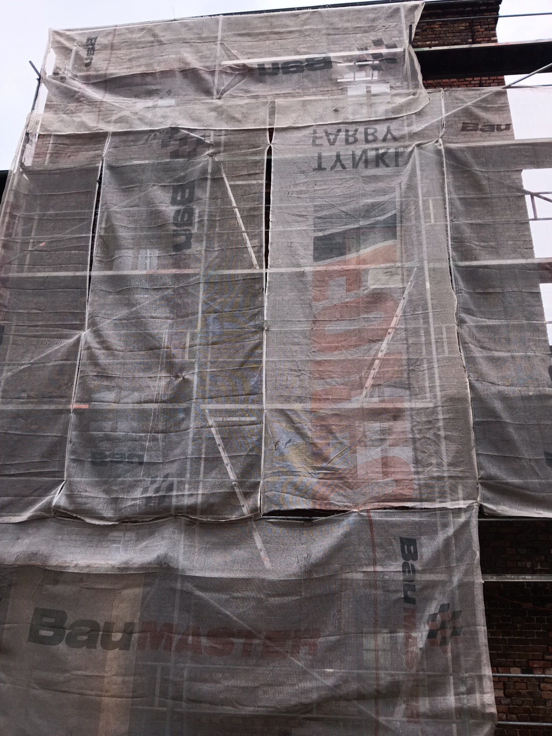 Budynek w trakcie prac elewacyjnych, osłonięty siatką ochronną z widocznym rusztowaniem i fragmentem ceglanej ściany. Ujęcie z dołu, pochmurne niebo w tle.