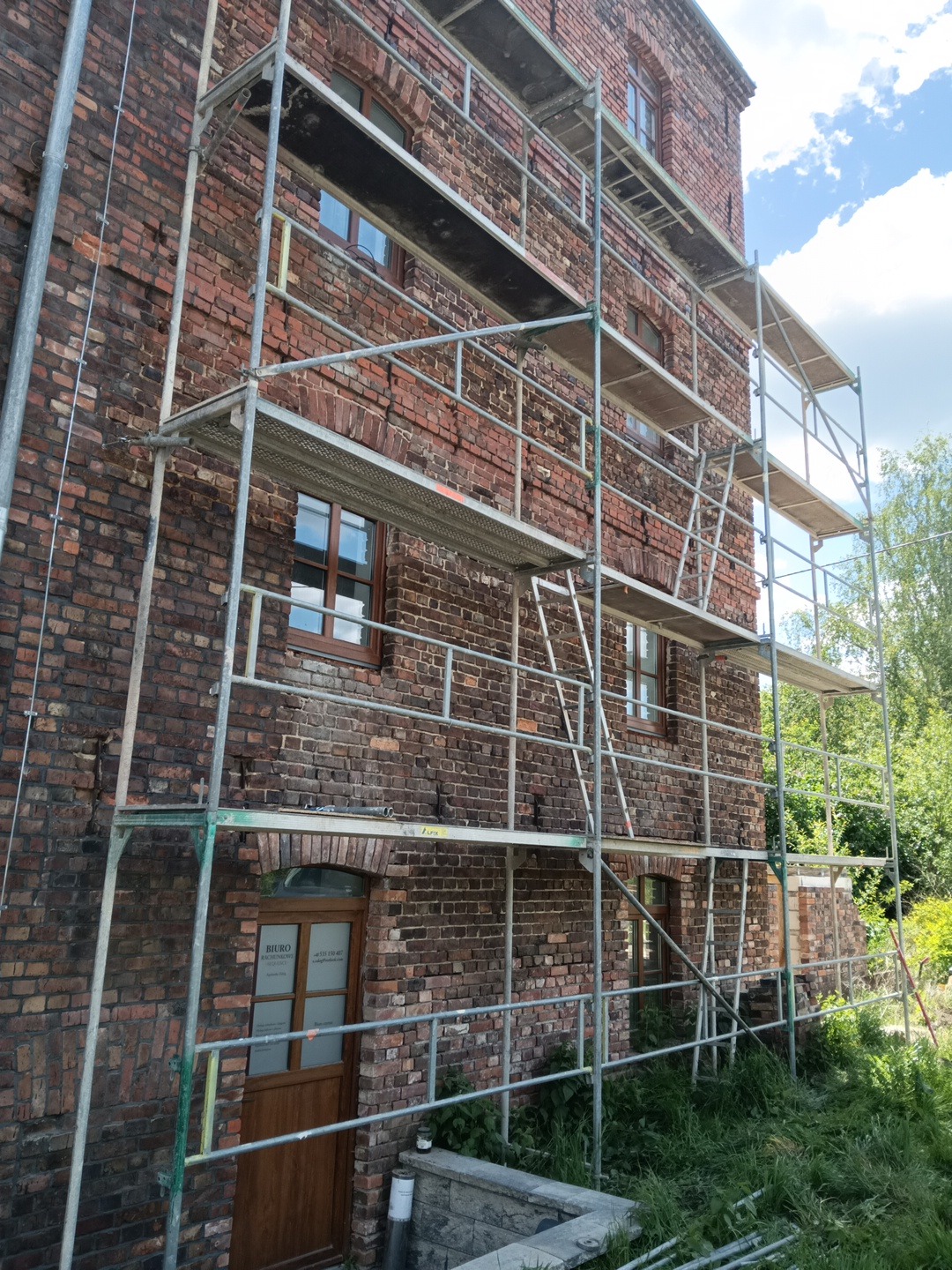 Budynek z czerwonej cegły w trakcie renowacji, otoczony rusztowaniami. Widoczne okna z drewnianymi ramami i drzwi wejściowe. Prace na wysokościach w słoneczny dzień.