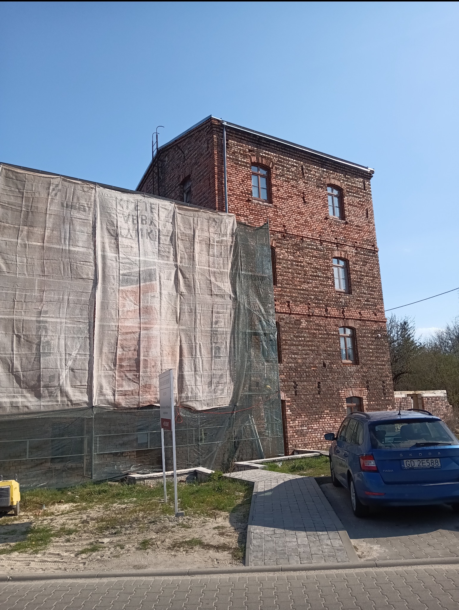 Ceglany budynek w trakcie renowacji, osłonięty siatką ochronną. Widoczny fragment elewacji z oknami i zaparkowany samochód osobowy. Słoneczny dzień.