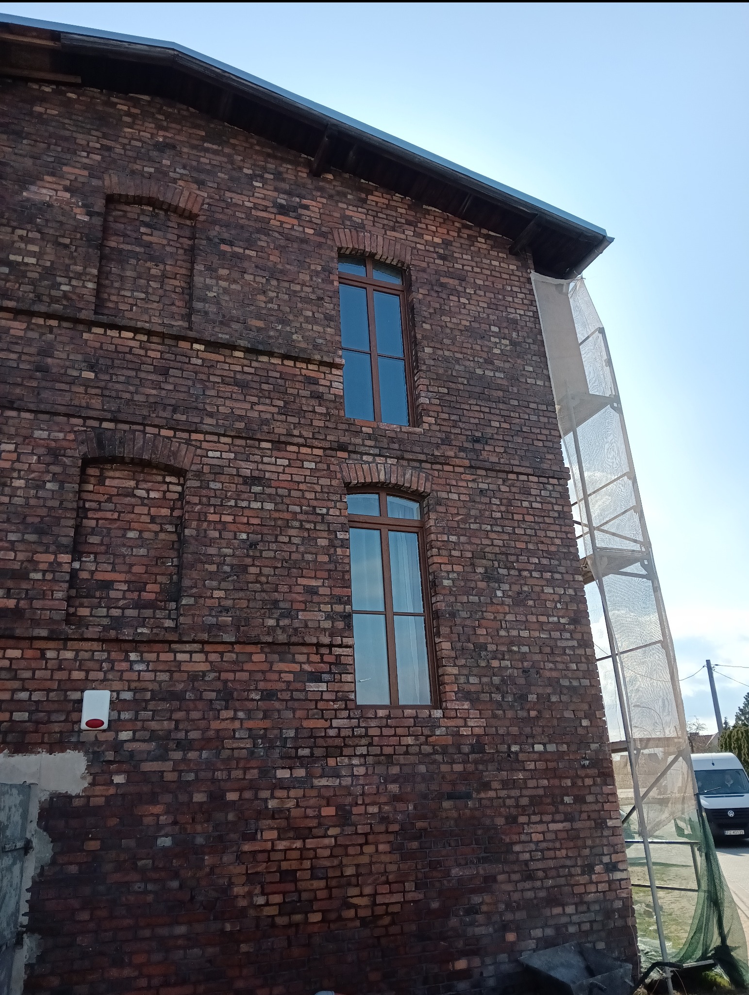 Ceglana elewacja budynku z nowymi oknami PCV i rusztowaniem. Widoczne ślady prac murarskich przy fundamencie i alarm. Ujęcie z dołu, błękitne niebo w tle.