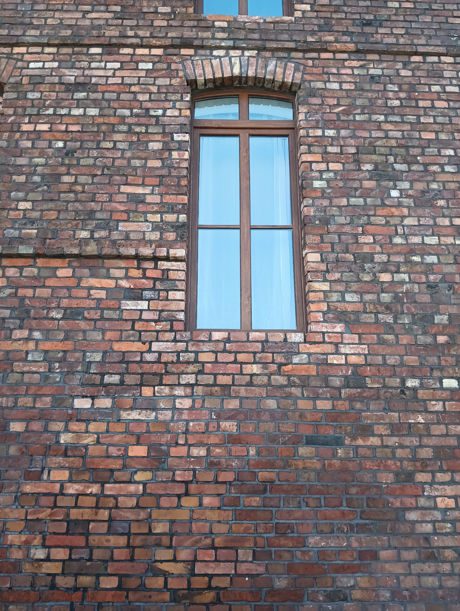 Ceglana ściana budynku z oknem PCV o brązowych ramach. Widoczne detale muru i refleksy światła na szybie. Charakterystyczna architektura.