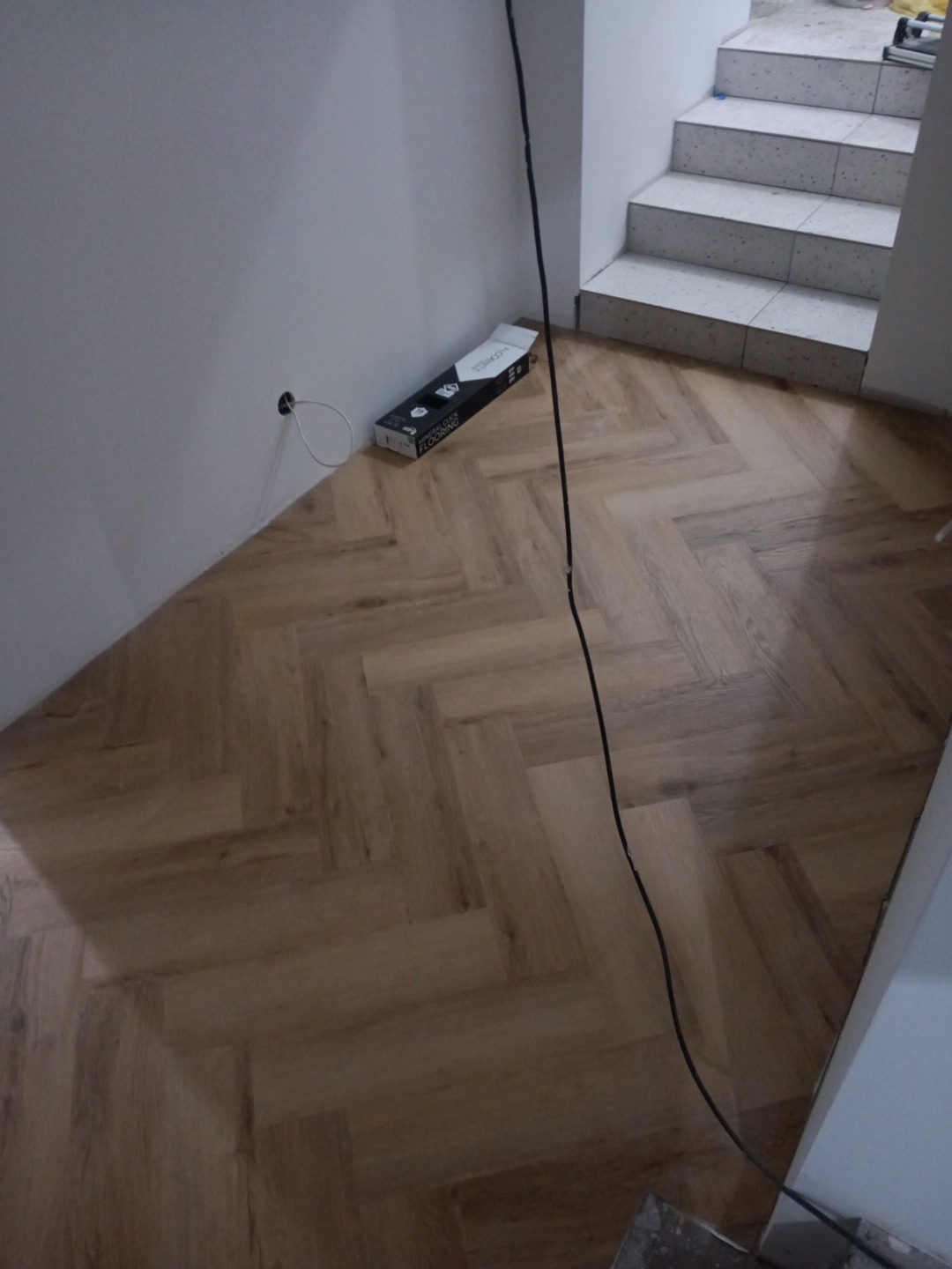 Ułożona podłoga z paneli w jodełkę, widoczne schody z terakoty i karton paneli przy ścianie. Kabel zwisa z góry. Jasne wnętrze w trakcie prac wykończeniowych.