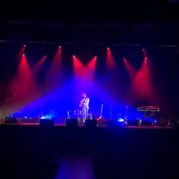 Koncert z gitarzystą na scenie oświetlonej czerwonymi i niebieskimi reflektorami, widok z widowni.
