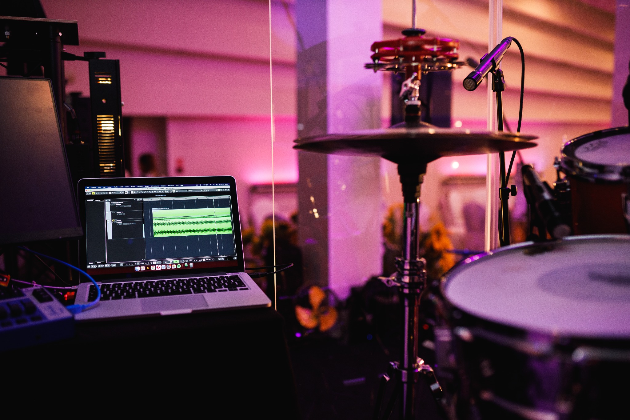 Stanowisko pracy realizatora dźwięku z laptopem wyświetlającym program do obróbki audio, mikrofonem i zestawem perkusyjnym w tle, oświetlone fioletowym światłem.