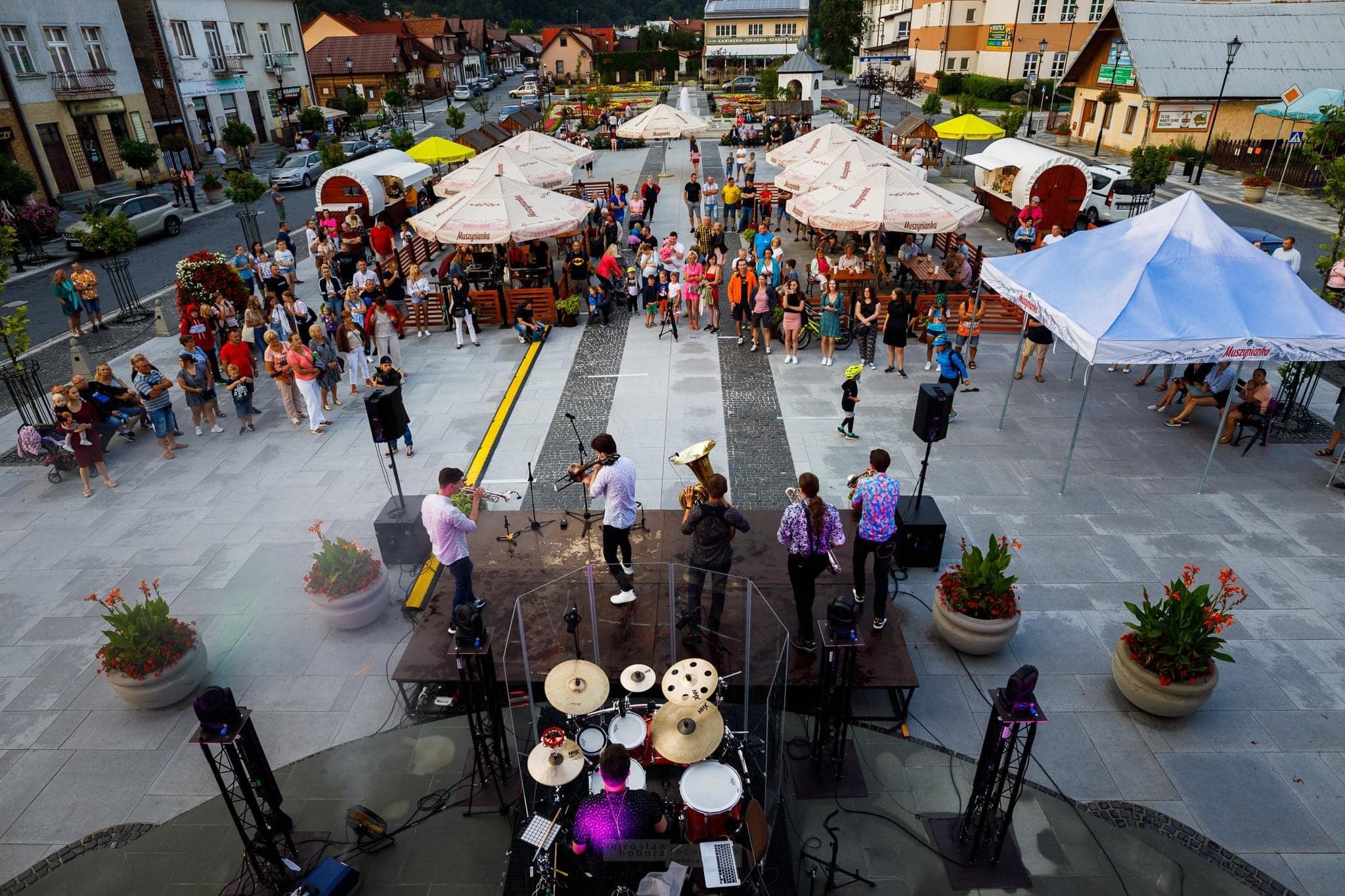 Zespół muzyczny gra na scenie podczas plenerowego wydarzenia na rynku miejskim z parasolami i food truckami w tle.