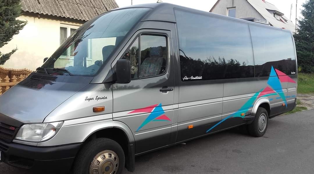 Srebrny bus Mercedes-Benz Super Sprinter z przyciemnianymi szybami i kolorowymi naklejkami w kształcie trójkątów, zaparkowany na tle domu.