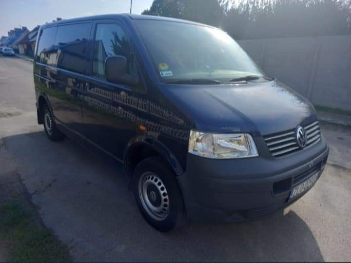 Niebieski Volkswagen Transporter T5 zaparkowany na asfaltowej drodze w słoneczny dzień, widoczne budynki w tle.