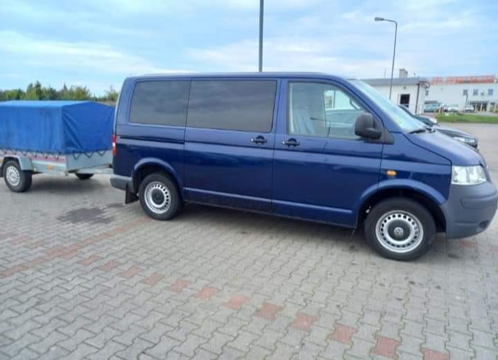 Niebieski van z przyczepką z plandeką na parkingu z kostki brukowej.