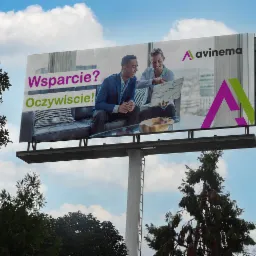 Billboard z dwoma uśmiechniętymi mężczyznami siedzącymi na kanapie, omawiającymi projekt na laptopie, z napisem 'Wsparcie? Oczywiście!' i logo firmy avinema na tle błękitnego nieba z chmurami.
