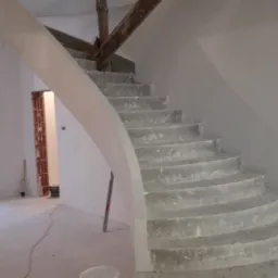 Wnętrze domu w trakcie prac wykończeniowych: spiralne schody w surowym stanie po nałożeniu pierwszej warstwy gładzi, widoczne drewniane elementy konstrukcyjne, pomieszczenie przed malowaniem...