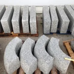 Przygotowane do montażu elementy z jasnoszarego, ziarnistego kamienia, niekt&oacute;re proste, inne łukowe, ułożone na paletach w magazynie.