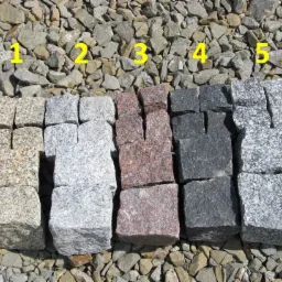 Przykładowe kostki brukowe z różnych rodzajów kamienia (granit, bazalt) ułożone na żwirze, prezentujące różnorodność kolorów i faktur.