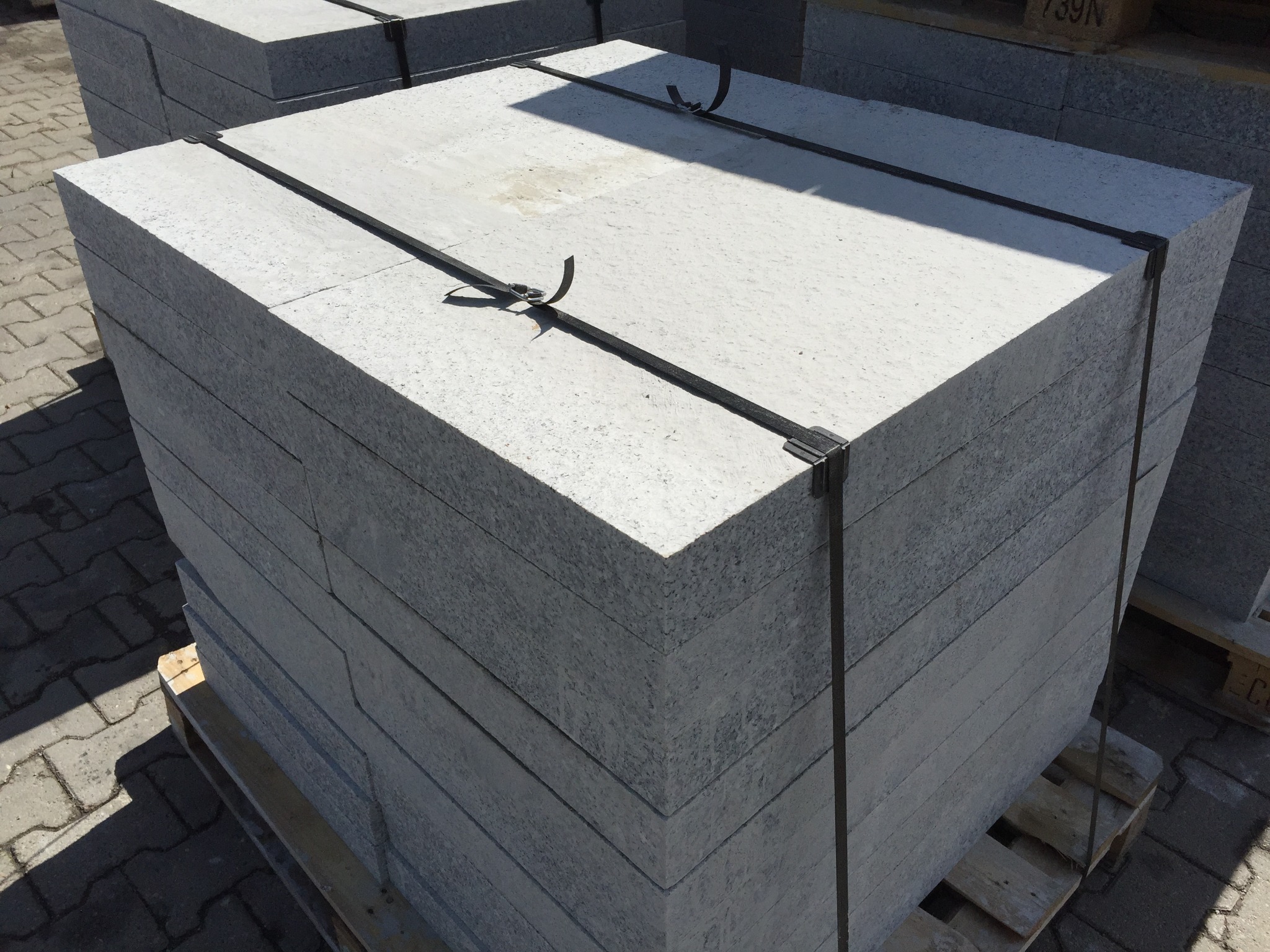 Stos szarych, kwadratowych płyt kamiennych na drewnianej palecie, zabezpieczonych czarną taśmą, w jasnym oświetleniu.