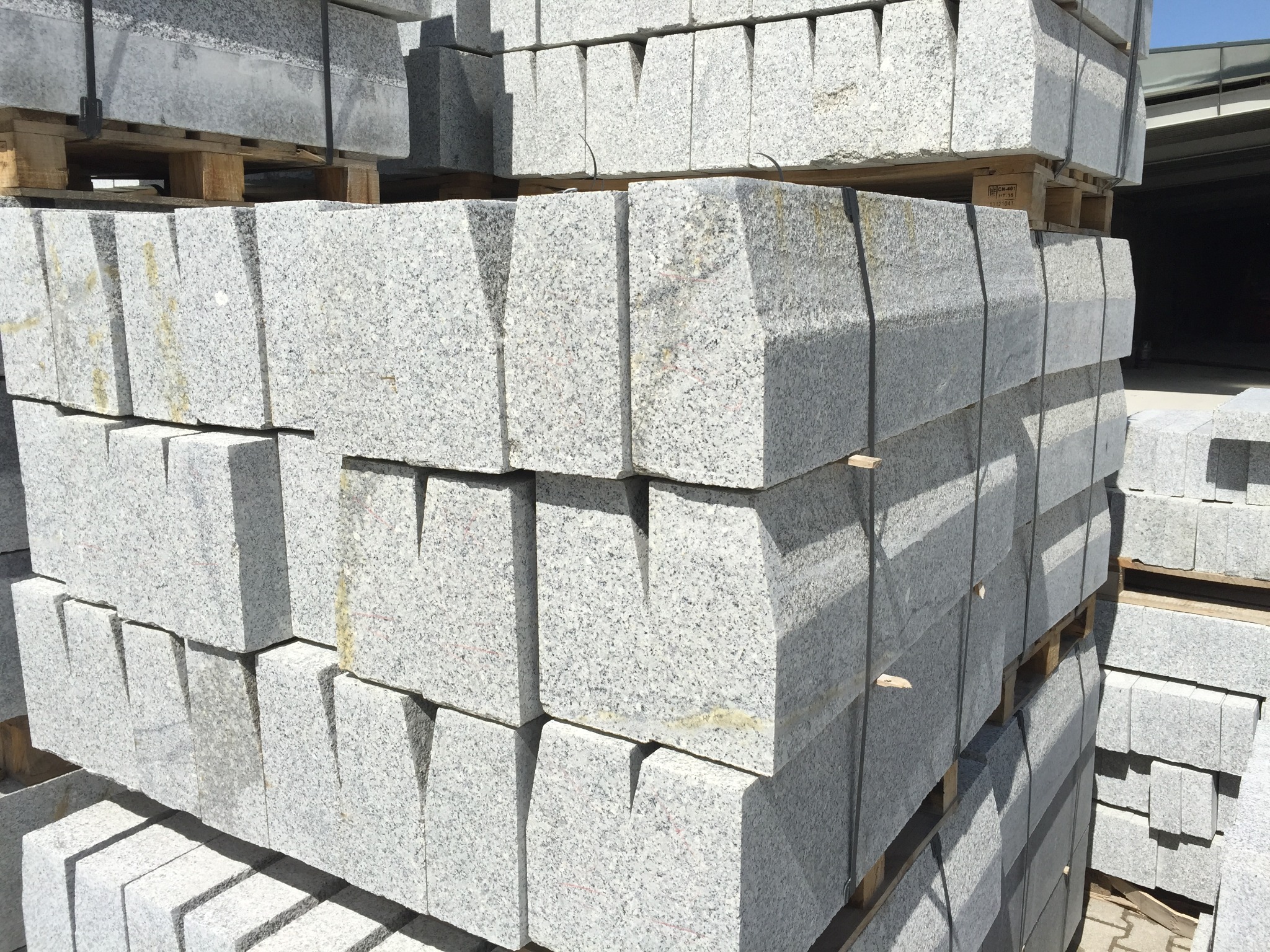 Szare, granitowe elementy krawężnikowe z wycięciami, ułożone na paletach, gotowe do transportu.