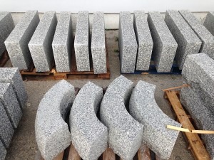 Przygotowane do montażu elementy z jasnoszarego, ziarnistego kamienia, niektóre proste, inne łukowe, ułożone na paletach w magazynie.