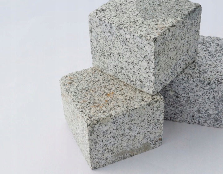 Trzy kostki z szarego, nakrapianego kamienia ułożone jedna na drugiej na białym tle. Widoczna szorstka tekstura kamienia.