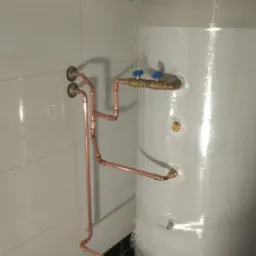 Biały zasobnik ciepłej wody owinięty folią ochronną, podłączony miedzianymi rurami w nowej instalacji, widok z boku, na podłodze płytki w kolorze grafitowym.