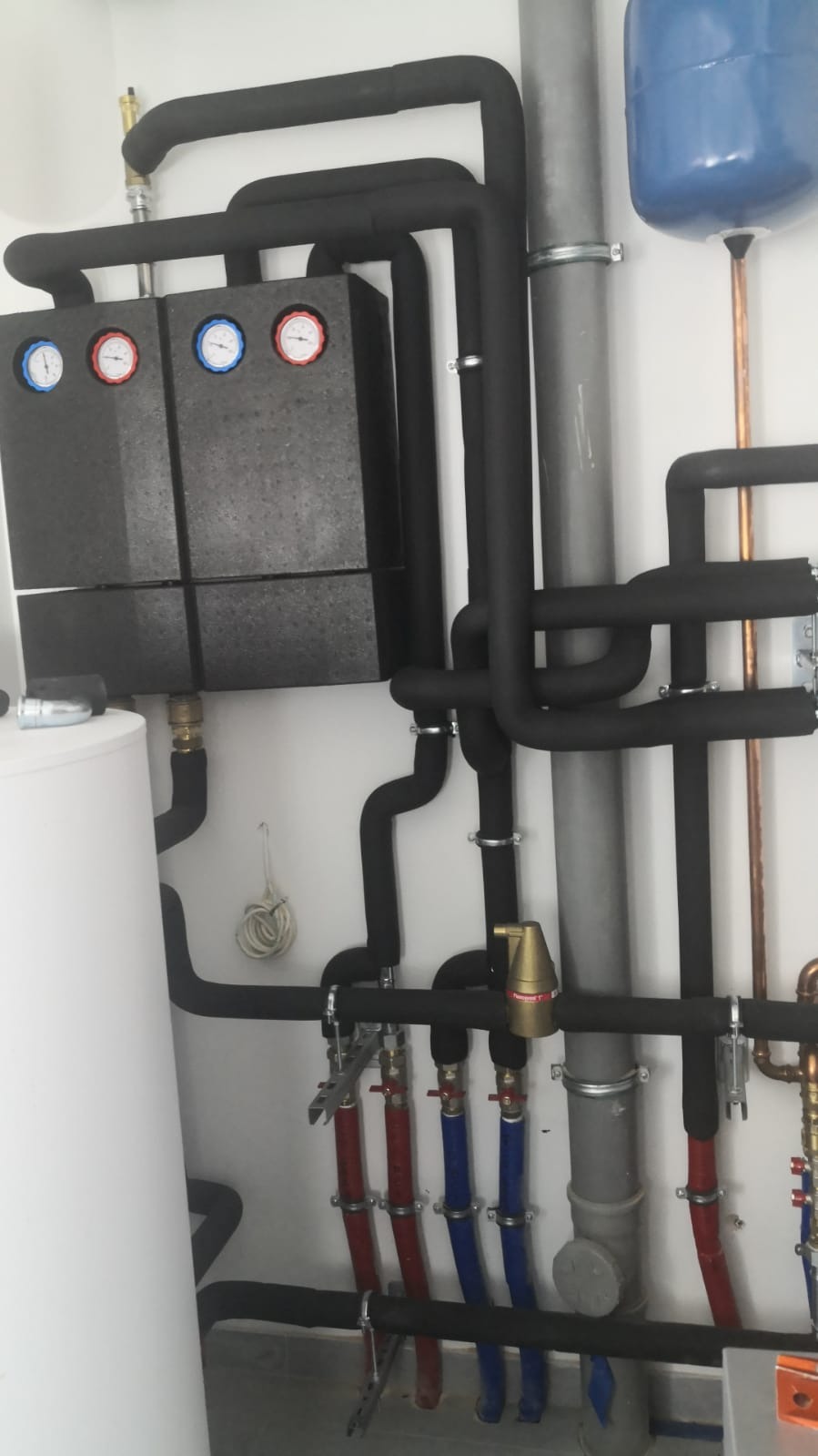 Zamknięty system grzewczy z czarnymi rurami izolowanymi termicznie, manometrami z czerwonymi i niebieskimi wskazówkami, oraz białym zbiornikiem wyrównawczym; widoczne zawory i elementy instalacji...