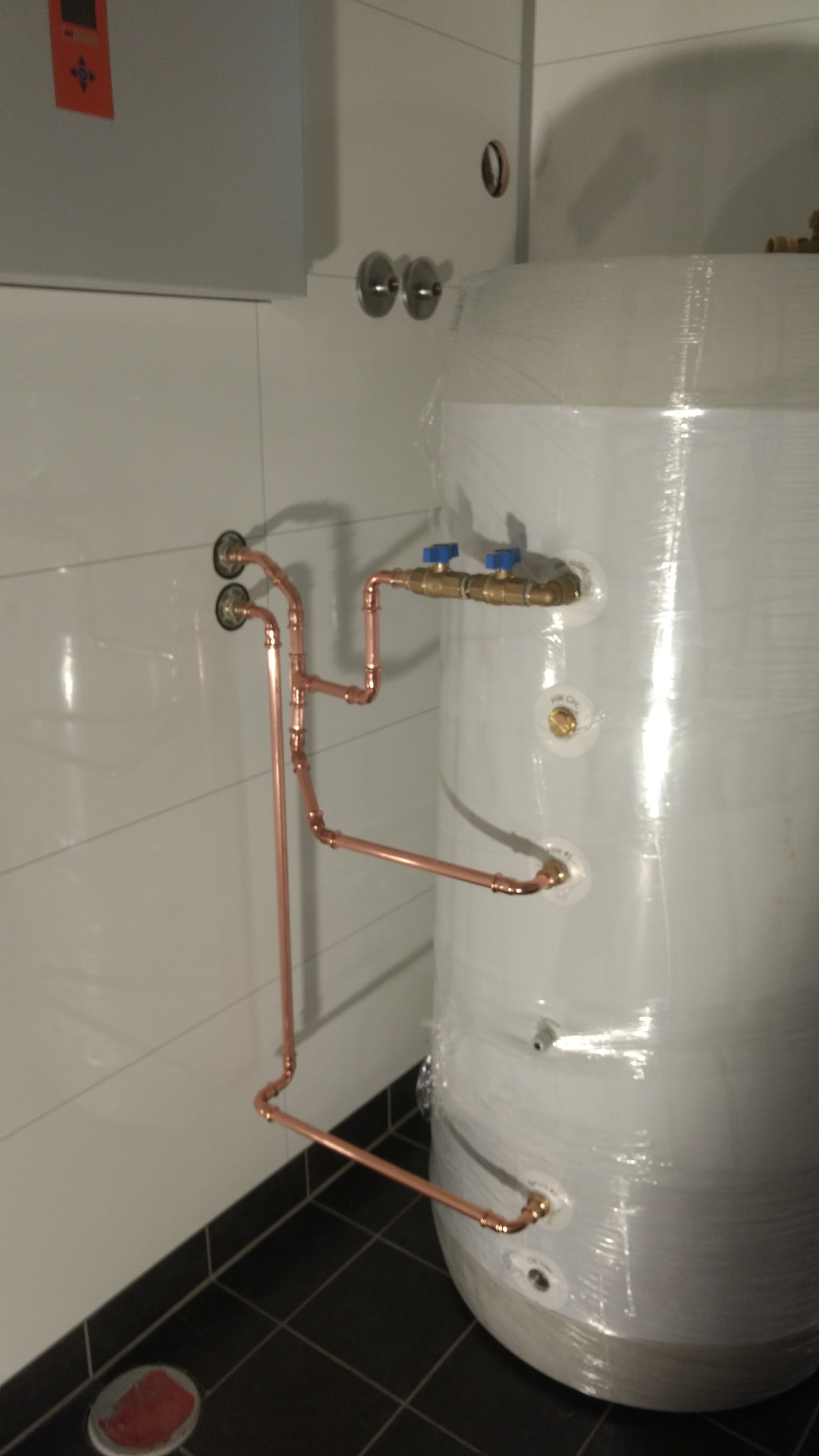 Biały zasobnik ciepłej wody owinięty folią ochronną, podłączony miedzianymi rurami w nowej instalacji, widok z boku, na podłodze płytki w kolorze grafitowym.