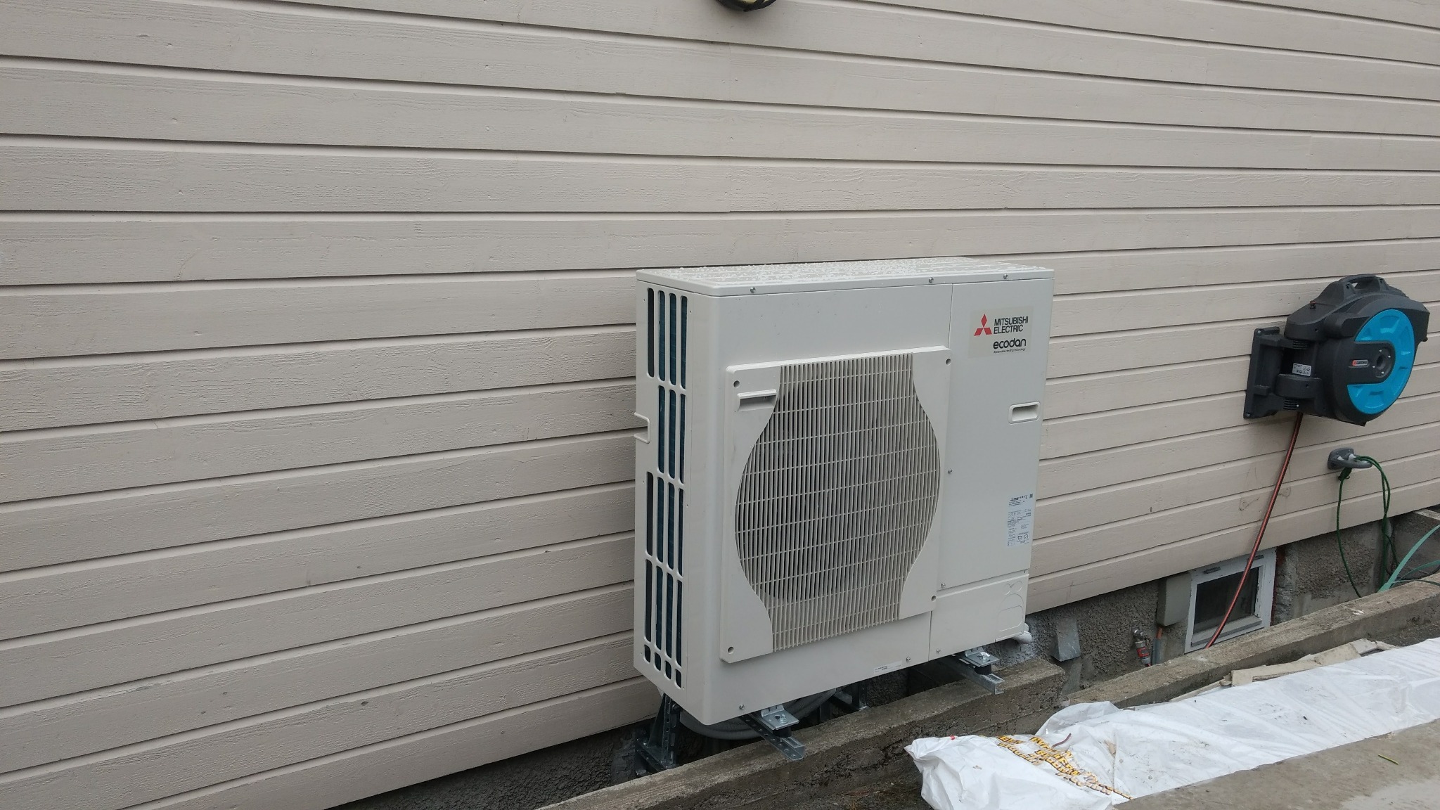 Biała jednostka zewnętrzna pompy ciepła Mitsubishi Electric Ecodan zamontowana na metalowej konstrukcji przy ścianie budynku z drewnianą elewacją, obok zwijadło węża ogrodowego.