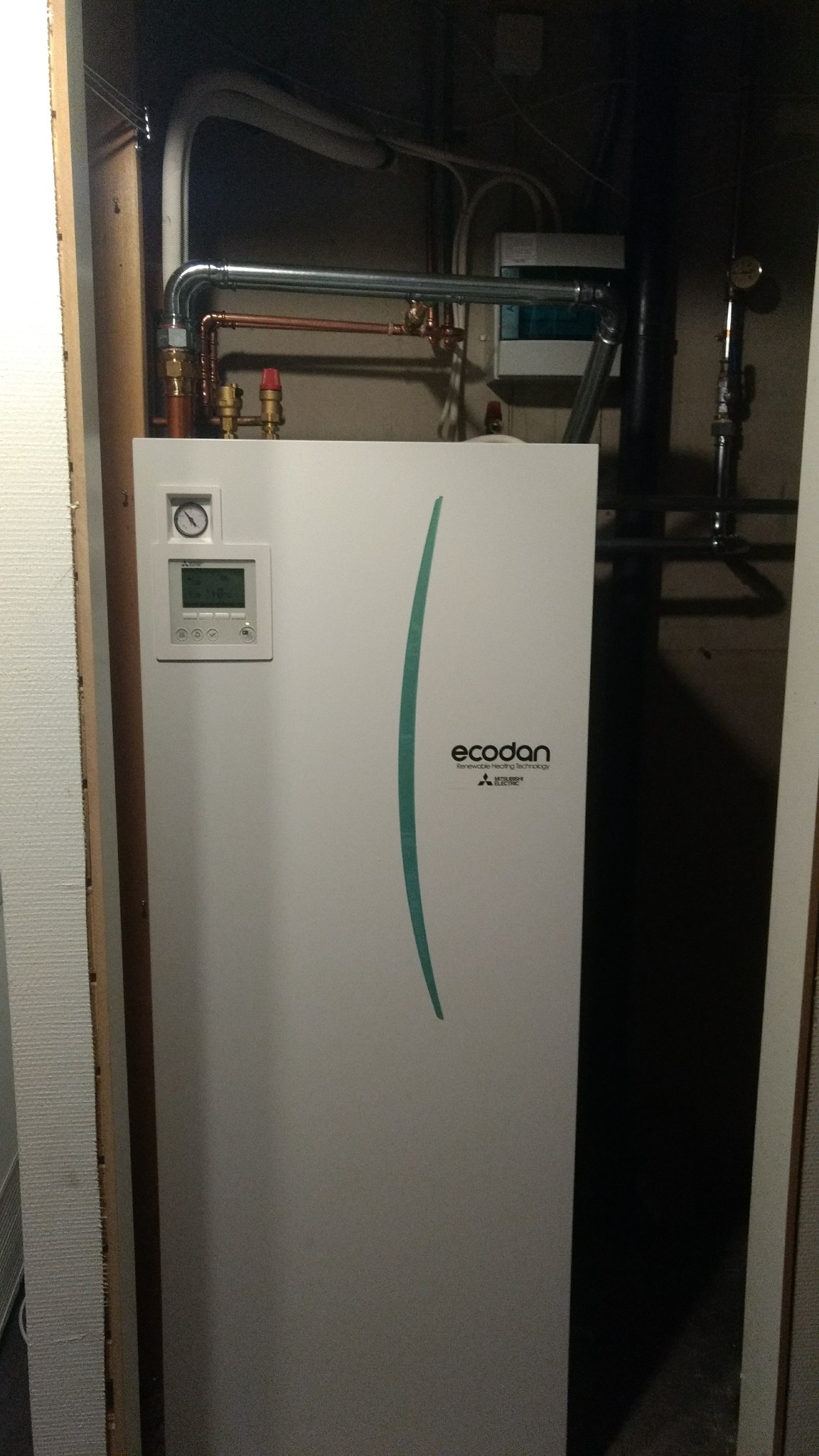 Biała jednostka Ecodan zamontowana w ciasnej przestrzeni z widocznymi rurami miedzianymi i elementami instalacji.