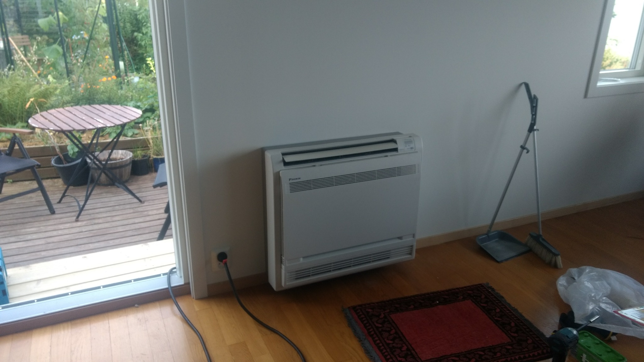 Biała, podłogowa klimatyzacja Daikin zamontowana przy ścianie w pokoju, widoczny kabel zasilający, w tle przez otwarte drzwi taras z meblami ogrodowymi, obok klimatyzacji stoi zmiotka z szufelką.