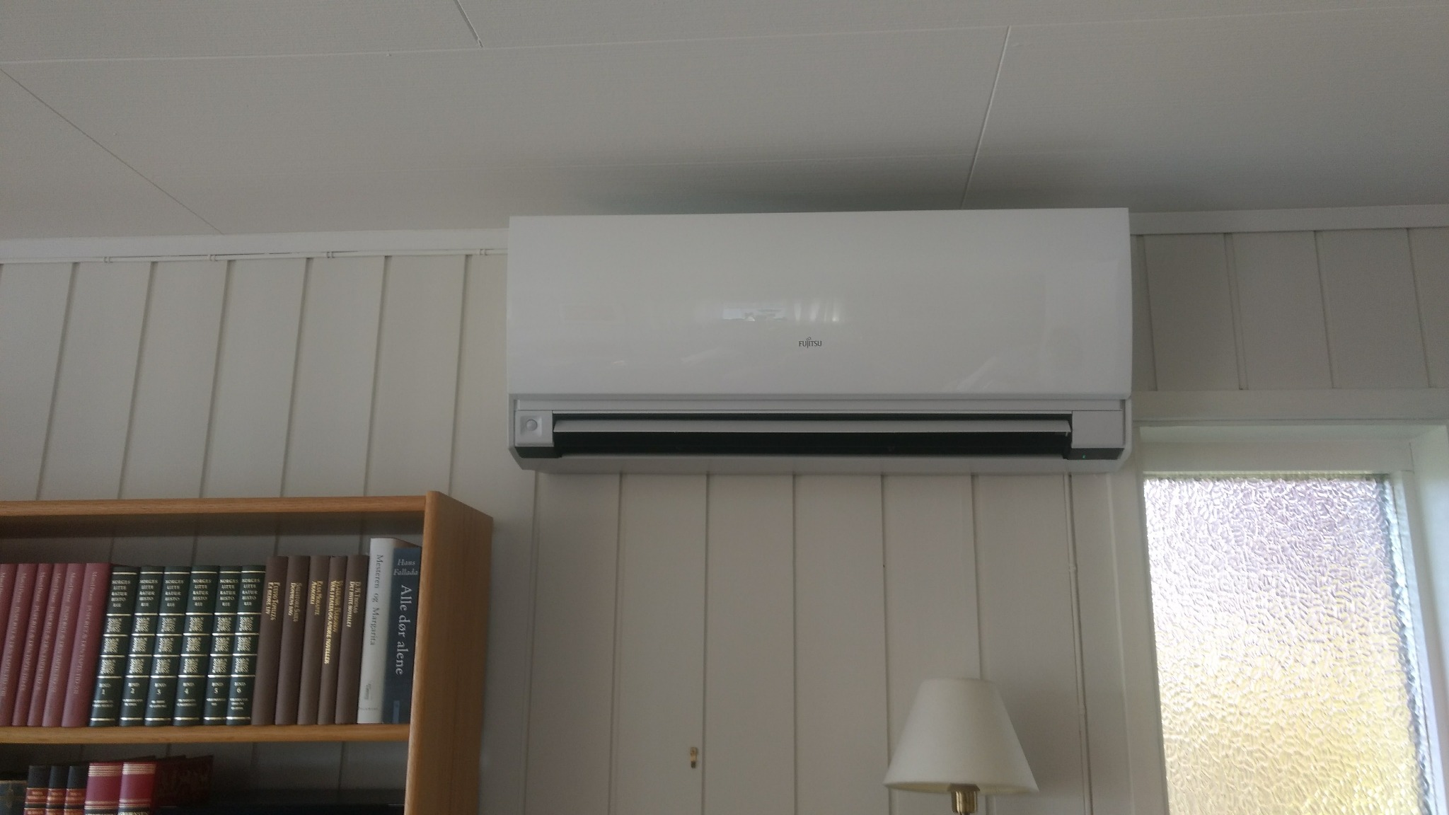 Biała jednostka klimatyzacyjna Fujitsu zamontowana na ścianie z boazerią, obok okna z matową szybą, z widocznym fragmentem regału z książkami i lampką nocną.