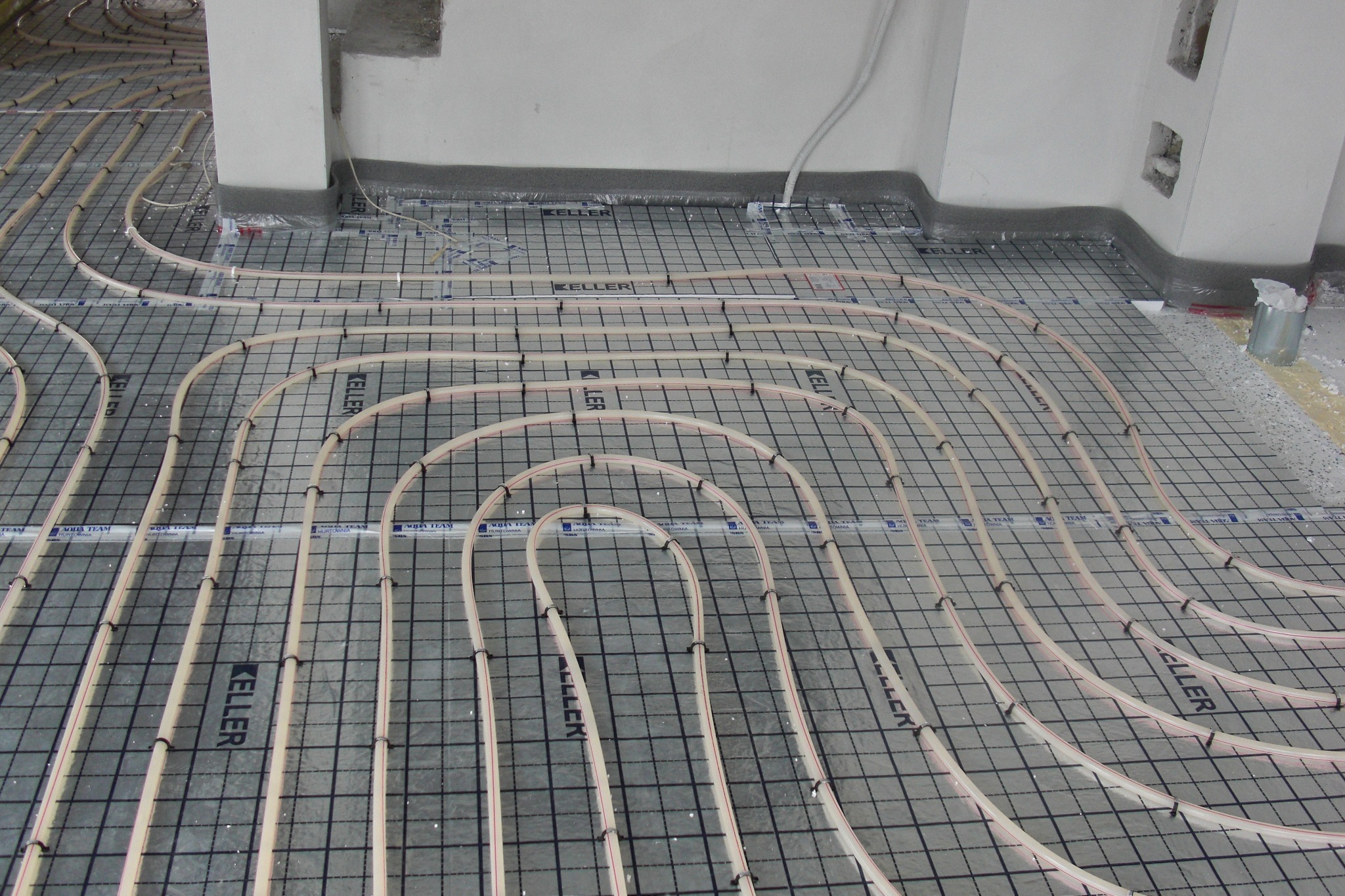 Rury grzewcze ułożone na siatce w trakcie instalacji ogrzewania podłogowego, widoczne czarne klipsy mocujące i izolacja przy ścianie.