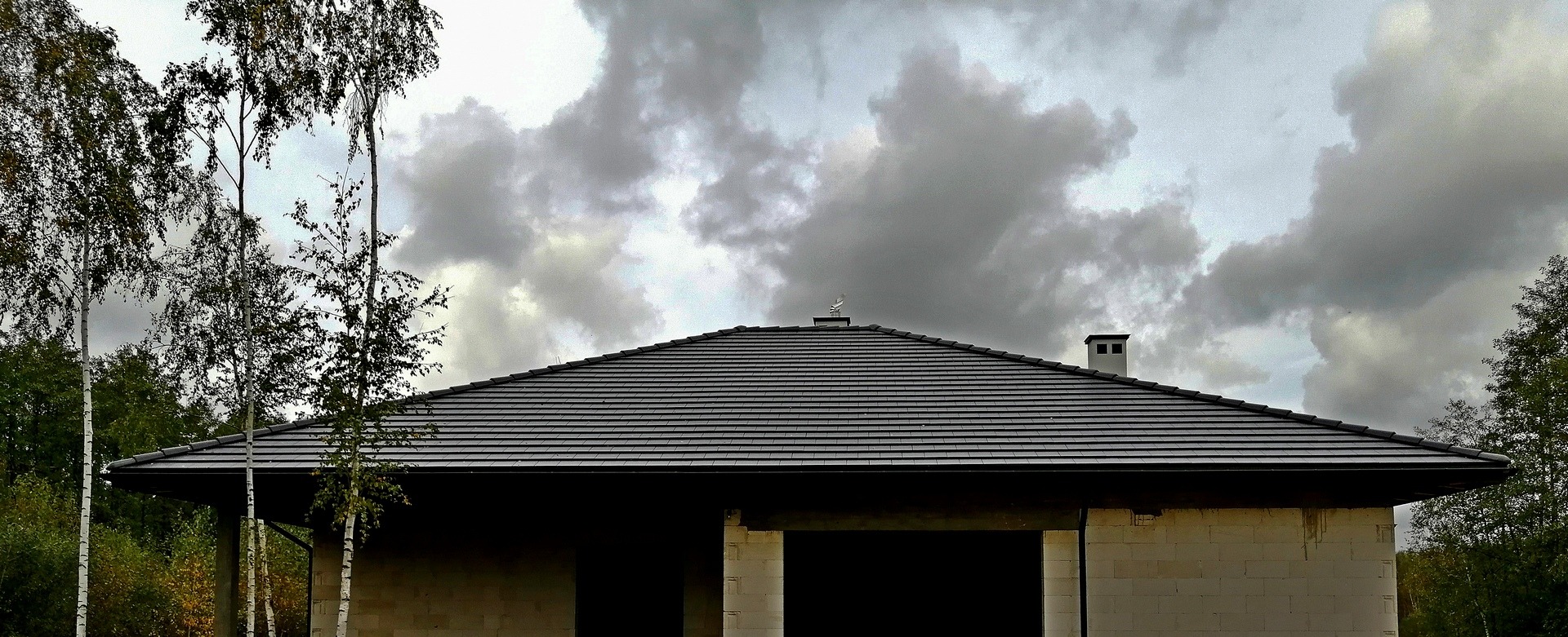 Dachówka cementowa Braas Tegalit Star na nowo wybudowanym domu jednorodzinnym z surowymi ścianami, widoczne brzozy i pochmurne niebo.