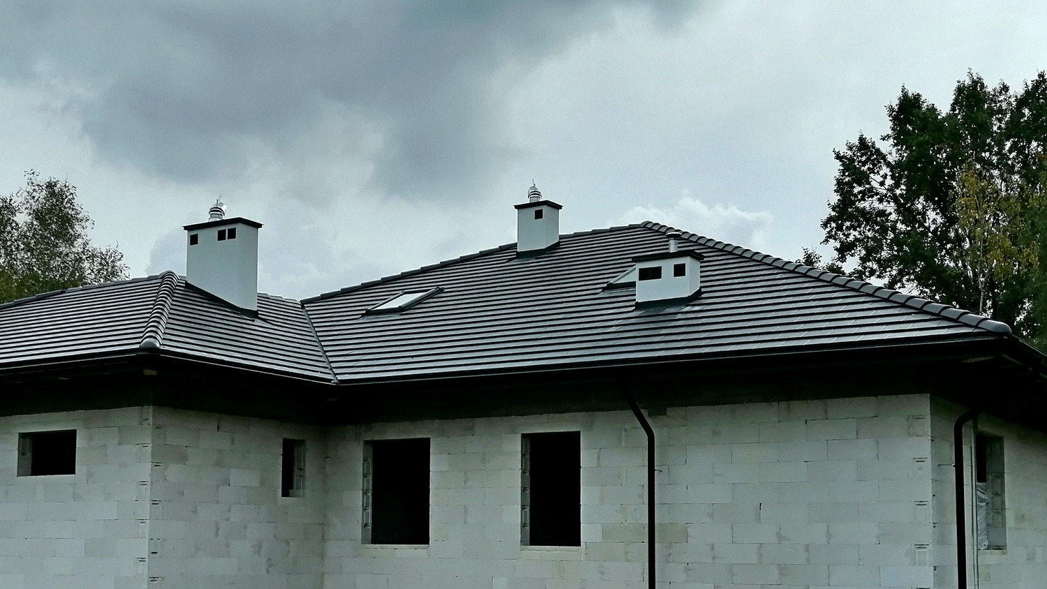 Dom w trakcie budowy, widoczny dach pokryty ciemną dachówką cementową Braas Tegalit Star, białe kominy z metalowymi zakończeniami, ściany z białego betonu komórkowego z otworami okiennymi...