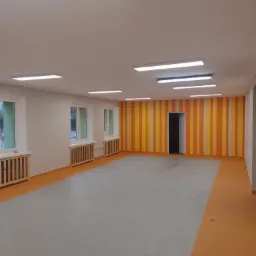 Wnętrze nowo wykończonego pomieszczenia z szarą podłogą, pomarańczowymi akcentami, grzejnikami pod oknami i ścianą w pionowe pasy żółto-pomarańczowe