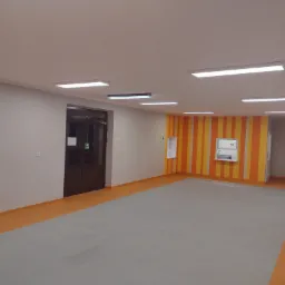 Wnętrze nowo wykończonego pomieszczenia z szarą podłogą i pomarańczowymi akcentami, widoczne drzwi wejściowe, ściana w pasy oraz oświetlenie sufitowe.