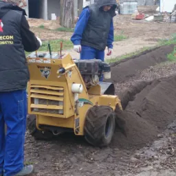 Żółta glebogryzarka Vermeer RT100 w trakcie wykonywania wykopu pod kable elektryczne, dwóch pracowników w odzieży roboczej nadzoruje prace, w tle teren budowy.